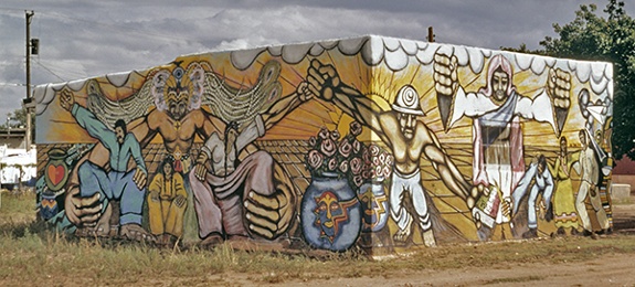 Mural by Artes Guadalupaños de Aztlán titled Saint Francis Drive Mural (1972). View of two sides of a building covered with mural featuring people, Quetzalcoatl, vases of flowers against a background of sun rays across fields.