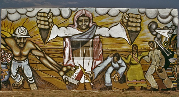 Mural by Artes Guadalupaños de Aztlán titled Saint Francis Drive Mural (1972). Central figure in white with a figure in a hard hat to the left and figure holding a pencil to the right with clouds, yellow sun, and fields behind.