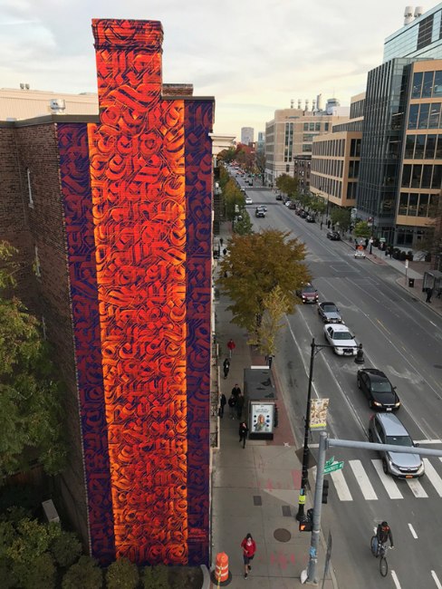 Mural by Sneha Shrestha (IMAGINE) titled For Cambridge with Love from Nepal (2019). Side of a five-story building with mural of Nepali calligraphy in oranges and violet-blues.